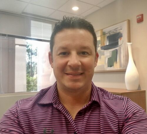 Justin Gilstrap, a Caucasian male in his 40s, sits at his desk and wears a purple polo shirt